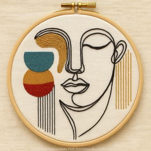 May include: An abstract embroidery art piece featuring a minimalist face design. The artwork uses black thread for the face outline and colorful accents of teal, gold, and red. The piece is set in a wooden embroidery hoop.