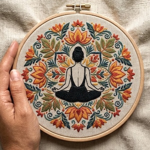 Puede incluir: Un bastidor de bordado circular de madera que contiene una tela beige con un diseño cosido a mano. El bordado presenta una figura central con ropa negra en posición de loto, rodeada por un patrón floral simétrico tipo mandala.