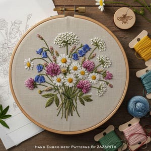 Puede incluir: Un diseño floral bordado a mano en un bastidor de madera. El bordado presenta un ramo de margaritas, tréboles y campanillas en tonos blancos, rosas y azules. El texto "Hand Embroidery Patterns By Zazarita" está en la parte inferior.