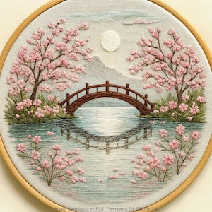 Puede incluir: Obra de arte bordada a mano en un aro de madera. La escena representa un paisaje sereno con un puente de madera sobre el agua, cerezos en flor y una luna llena. La paleta de colores incluye tonos de rosa, verde, marrón y azul.