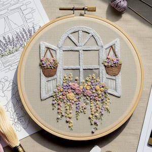May include: Embroidered hoop art featuring a white window with open shutters and hanging baskets. The window is adorned with colorful flowers in shades of pink, yellow, and purple. The embroidery is set against a neutral fabric background.