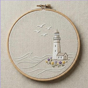 May include: A circular wooden embroidery hoop containing a beige fabric panel with a hand-stitched design of a white lighthouse, stylized ocean waves, four flying birds, and a small cluster of purple and yellow flowers at the base.