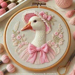 May include: An embroidered goose wearing a pink bonnet and bow, set in a wooden hoop. The artwork features detailed stitching in white, pink, and peach thread, with floral accents and the text "Hand Embroidery Patterns By Zazarita".