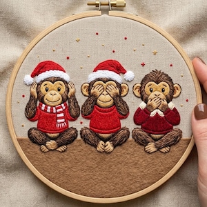May include: Embroidered artwork of three monkeys in a wooden hoop. Each monkey wears a red sweater and Santa hat, with one covering its ears, one its eyes, and one its mouth. The background is beige.