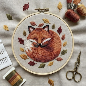 May include: Embroidered artwork in a wooden hoop, depicting a sleeping fox in orange and white, encircled by autumn leaves in various colours. Additional embroidery supplies, including scissors and thread, are also visible.