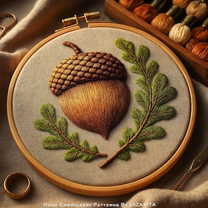 Op de afbeelding: Geborduurde eikel en eikenblad ontwerp in een houten hoepel. De eikel is bruin met een getextureerde dop en de bladeren zijn groen. De tekst "HAND EMBROIDERY PATTERNS BY ZAZARITA" staat onderaan de afbeelding. De achtergrond is een neutrale stof.