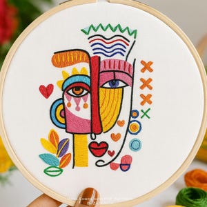 May include: An abstract face embroidered on white fabric, framed in a wooden hoop. The design uses bold lines and shapes in pink, yellow, orange, blue, and red. Includes heart and leaf accents, creating a modern art piece.