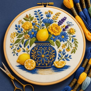 May include: Embroidered hoop art featuring a blue vase with yellow lemons and flowers. The embroidery includes yellow roses, blue flowers, and green leaves. Yellow and blue embroidery floss and gold scissors are also in the image. The artwork is set against a dark blue background.