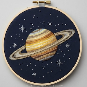 May include: Hand-stitched embroidery art of Saturn, featuring a planet with yellow, brown, and white bands and rings, set against a dark blue background with embroidered stars and silver beads. The artwork is displayed in a wooden embroidery hoop.