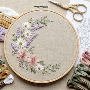 May include: A floral embroidery hoop featuring a crescent design of pink, white, and lavender flowers with green leaves. The embroidery is set in a wooden hoop, with spools of thread and gold scissors nearby. The craft project is on a neutral linen fabric.