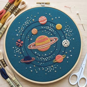 May include: Hand-stitched embroidery art of a solar system on teal fabric. Planets in shades of orange, yellow, and blue are arranged around a central planet with rings. White star details and a wooden hoop complete the design. Embroidery floss and scissors are visible.