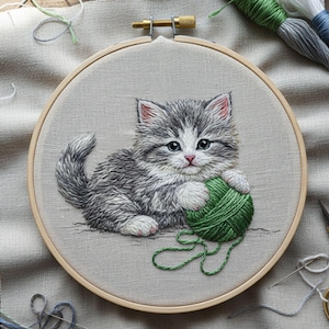 Puede incluir: Obra bordada de un gatito gris y blanco con ojos azules, sosteniendo una bola de hilo verde. El bordado está en un bastidor de madera, con varios hilos de colores y una aguja visibles en el fondo.