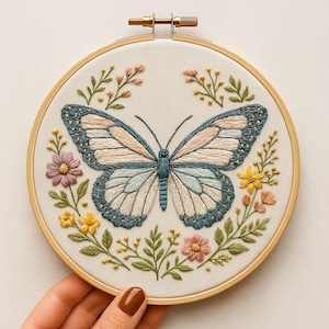 May include: Embroidered butterfly artwork in a wooden hoop. The butterfly features blue, pink, and cream wings with detailed stitching. The design is encircled by embroidered flowers and foliage in pink, yellow, and green hues.