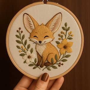 May include: Embroidered hoop art featuring a smiling fox with yellow and white flowers. The fox is sitting on a bed of green leaves.
