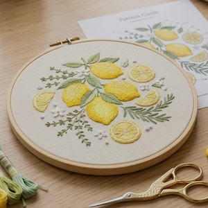 Könnte beinhalten: Fertiggestickter Stickrahmen mit einem Zitronen-Design. Das Design zeigt gelbe Zitronen, grüne Blätter und weiße Blüten. Im Hintergrund ist eine Musteranleitung zu sehen. Stickgarn und eine goldene Schere sind ebenfalls vorhanden.