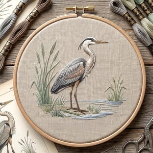 May include: An embroidered heron in a wooden hoop, with reeds and water details. The heron has grey and brown plumage. Embroidery threads in various shades are arranged around the hoop. The hoop is set on a wooden surface.