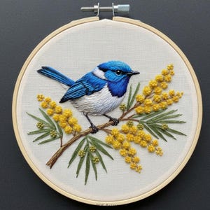 May include: Hand-stitched embroidery of a blue and white bird on a branch with yellow flowers and green foliage. The artwork is displayed in a round wooden hoop, set against a white fabric backdrop.