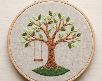 May include: Embroidered tree art in a wooden hoop. The tree features a brown trunk and branches with green leaves. A small wooden swing hangs from a branch. The background is a light tan fabric, and the hoop is secured with a brass screw.