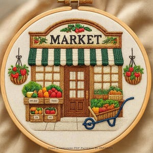 May include: Embroidered hoop art featuring a market scene. The design includes a wooden market facade with a green and white striped awning, baskets of produce, and the word "MARKET" above the entrance. Two hanging baskets of red tomatoes flank the market.