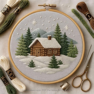 May include: Hand-embroidered winter scene in a wooden hoop. The artwork depicts a snow-covered cabin nestled among evergreen trees and mountains. Embroidery floss in shades of green, brown, and white surrounds the hoop, along with gold scissors and thread spools.