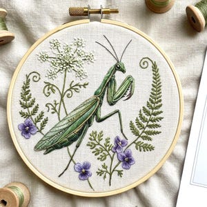 May include: Embroidered hoop art featuring a detailed green praying mantis, surrounded by white flowers, purple violets, and green foliage. The embroidery is set in a natural wood hoop, creating a nature-inspired piece.