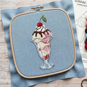 Puede incluir: Bordado de un sundae en un vaso, con helado de vainilla, fresa y chocolate, cubierto con crema batida y una cereza. El bordado está colocado en un aro de madera sobre una tela azul claro.