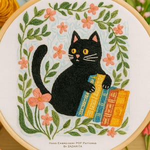 May include: Hand-embroidered hoop art featuring a black cat holding books, surrounded by a floral wreath of green leaves and peach-colored flowers. The cat has yellow eyes and white whiskers. The text at the bottom reads "Hand Embroidery PDF Patterns By Zazarita."