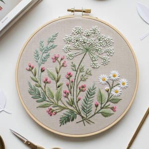 Puede incluir: Un bastidor de bordado con un diseño floral en hilo blanco, rosa y verde. El diseño incluye margaritas, pequeñas flores rosas y una flor blanca grande. La tela es de color beige claro y el bastidor es de madera, de unos 20 cm de diámetro.
