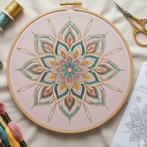 May include: A completed embroidery project in a wooden hoop. The design is a symmetrical floral pattern in shades of teal, pink, gold, and yellow on a light pink fabric. Gold embroidery thread outlines the design. Embroidery floss and scissors are visible.