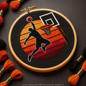 Puede incluir: Un bordado con temática de baloncesto en un aro de madera. El diseño muestra a un jugador haciendo un mate, silueteado contra un atardecer naranja y rojo. El aro está rodeado de hilos de bordar naranjas y negros, con un diámetro de unos 20 cm.