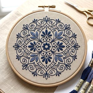May include: An embroidered mandala design in a wooden embroidery hoop. The design features a symmetrical pattern in shades of blue on a beige fabric. Embroidery floss and gold scissors are visible.