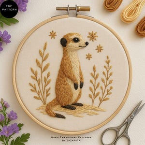 Puede incluir: Un suricato bordado a mano en un bastidor de madera. El suricato es de color marrón claro con detalles negros, rodeado de plantas bordadas y flores en forma de estrella. El texto "PDF PATTERN" y "HAND EMBROIDERY PATTERNS BY ZAZARITA" son visibles.