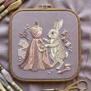 May include: An embroidered artwork featuring a rabbit measuring a dress on a mannequin. The dress is pink and purple, with lace and pearls. The artwork is in a square wooden frame, with spools of thread and scissors.