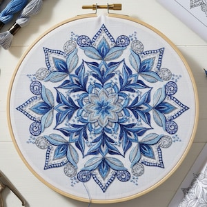 May include: A completed embroidery project featuring a detailed blue and gray floral mandala design. The embroidery is set in a wooden hoop, with various shades of blue and gray thread used to create the intricate pattern. Embroidery floss and a printed pattern are visible.