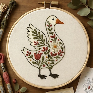 May include: A circular wooden embroidery hoop containing white fabric with a stitched design of a white goose. The goose is decorated with green leaves, red floral elements, and a single white daisy. The goose has a bright orange beak and black feet.