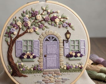 May include: Embroidered hoop art featuring a detailed scene of a house with a purple door, shutters, and a stone path. A tree with purple and cream-colored flowers arches over the house. The embroidery is set in a wooden hoop.