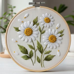 May include: A handcrafted embroidery hoop featuring three white daisies with yellow centers and green leaves. The floral design is set against a light-colored fabric, secured in a wooden hoop. Scattered white petals add a delicate touch.