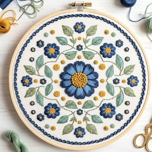 May include: An embroidered floral design on a white fabric stretched within a wooden hoop. The design features blue flowers with yellow centres, green leaves, and yellow dots. The hoop has a blue border and is ready for display.