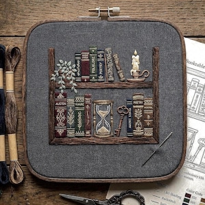 May include: Embroidered artwork of a bookshelf with books, a candle, and an hourglass. The books are labeled with titles like "Austen" and "Poetry." The piece is framed in a square hoop with a wooden frame and a gray background.