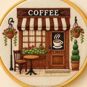 May include: Embroidered hoop art featuring a coffee shop scene. The shop has a brown and white striped awning, a door with a coffee cup graphic, and the word "COFFEE" above. Includes a small table, chair, and hanging flower baskets.