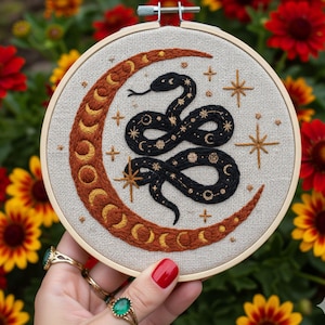 以下が含まれることがあります： 黒いヘビが三日月と絡み合った刺繍フープアート。月は温かみのある茶色で金色のアクセントがあり、ヘビには金色の星と月のディテールがあります。背景はニュートラルなリネン生地です。