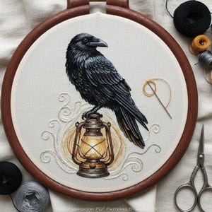 May include: Hand-embroidered raven on a lantern, in a wooden hoop. The raven is black with detailed feathering. The lantern glows with warm light, and the background is cream-coloured fabric. Embroidery thread and scissors are visible.