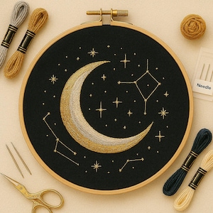 Puede incluir: Diseño celestial bordado sobre tela negra en un aro de madera. El diseño presenta una luna creciente dorada y blanca, rodeada de estrellas y constelaciones bordadas. También se ven hilos de bordar, agujas y tijeras.