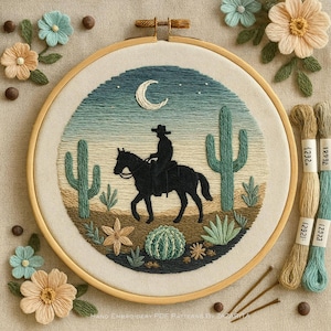 May include: Embroidered hoop art depicting a cowboy on horseback in a desert scene with cacti, a crescent moon, and stars. The artwork is in a wooden hoop, with floral embellishments and embroidery threads nearby.