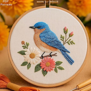 May include: Embroidered bluebird on a branch with flowers in a wooden embroidery hoop. The bird has blue and orange feathers, with white and coral flowers. The text "PDF PATTERN" is in the upper left corner.