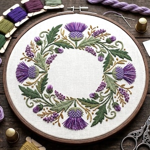 Könnte beinhalten: Ein runder Holzstickrahmen mit weißem Stoff, der ein handgesticktes Kranzmotiv zeigt. Die Stickerei besteht aus violetten Distelblüten, grünen gezackten Blättern und kleinen violetten Blütenclustern in kreisförmiger Anordnung.