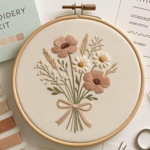 May include: An embroidery kit featuring a floral design. The artwork includes pink flowers, white daisies, and wheat-like stalks, all tied with a ribbon. The embroidery is set in a wooden hoop, with a kit box and thread spools visible.
