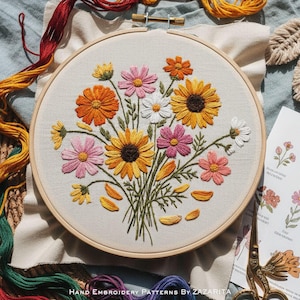 May include: A hand-embroidered floral design in a wooden hoop. The embroidery features a bouquet of colorful flowers, including orange, pink, yellow, and white daisies. The text "Hand Embroidery Patterns by Zazarita" is at the bottom.