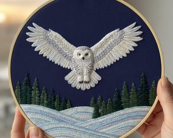 May include: Embroidered artwork featuring a snowy owl with outstretched wings, set against a dark blue background. The owl is white with gray accents, above a snowy landscape and evergreen trees. The artwork is framed in a wooden hoop.