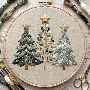 May include: A circular wooden embroidery hoop containing a linen fabric panel with three hand-embroidered Christmas trees. The trees feature shades of green, gold, and cream thread with gold star toppers and small circular accents throughout the design.